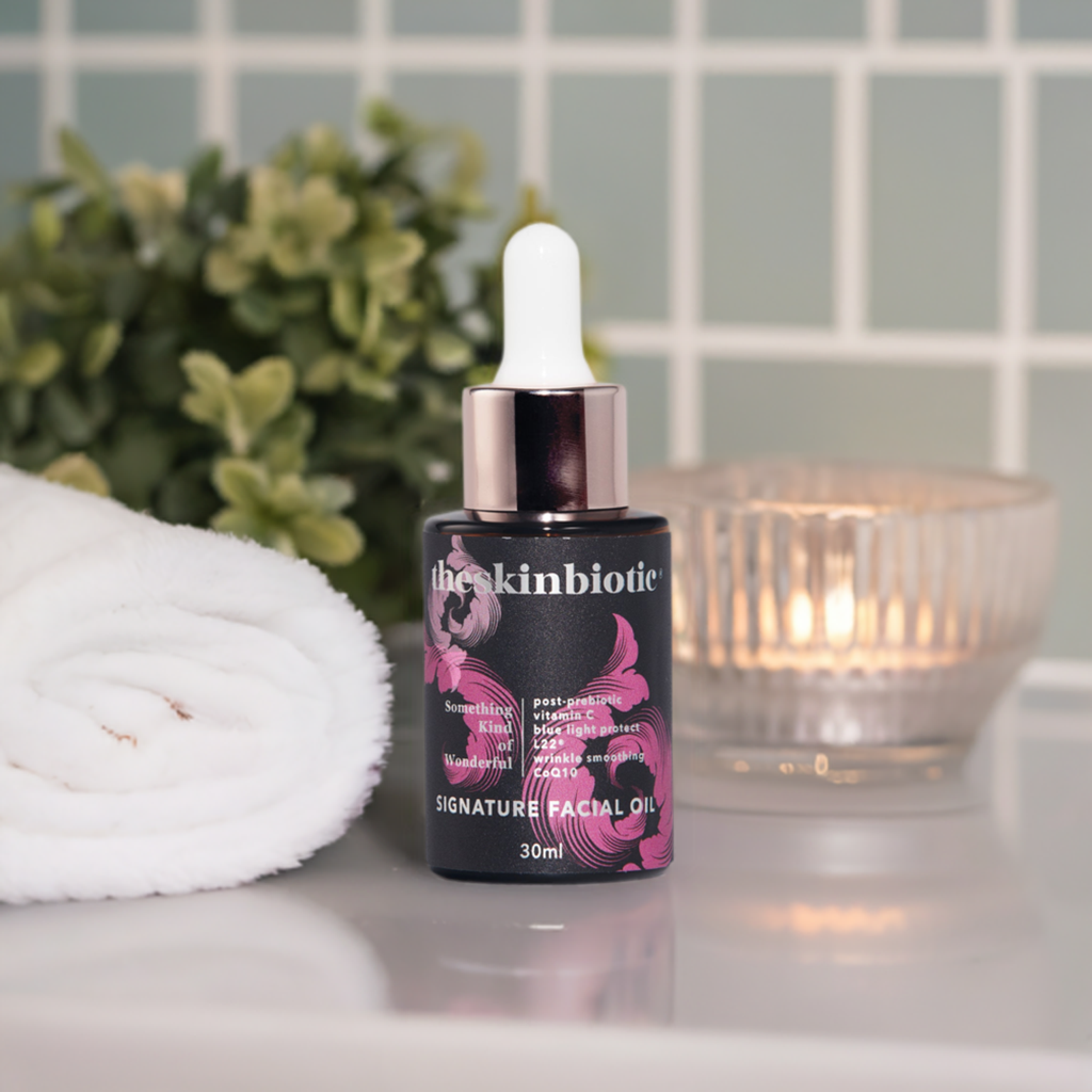 theSkinBiotic Signature Facial Oil picture of a bottle with a dropper on a bathroom bench with gentle candle lighting, a plant and white towel